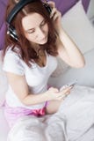 De aantrekkelijke jonge donkerbruine vrouw zit in de ochtend in haar bed met hoofdtelefoons en een smartphone en luistert binnen royalty-vrije stock fotografie De aantrekkelijke jonge donkerbruine vrouw zit in de ochtend in haar bed met hoofdtelefoons en een smartphone en luistert binnen royalty-vrije stock fotografie