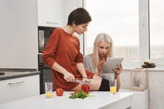 De fille de lecture de charger comprimé blond mignon avec son amie tandis qu'elle coupe des légumes, préparant la salade Chacun d photographie stock De fille de lecture de charger comprimé blond mignon avec son amie tandis qu'elle coupe des légumes, préparant la salade Chacun d photographie stock