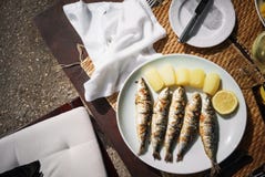 De gebraden smakelijke makreel, een plak van citroen en de gele aardappels liggen op een witte plaat, die zich op een houten lijs stock foto De gebraden smakelijke makreel, een plak van citroen en de gele aardappels liggen op een witte plaat, die zich op een houten lijs stock foto