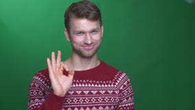 De gelukkige jonge donkerbruine mens in sweater maakt o.k. gebaar wordt verrukt op groene achtergrond stock videobeelden De gelukkige jonge donkerbruine mens in sweater maakt o.k. gebaar wordt verrukt op groene achtergrond stock videobeelden