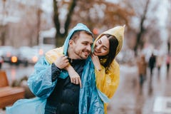 De gelukkige paar het houden van kerel en zijn meisje gekleed in regenjassen koesteren op de straat in de regen stock fotografie De gelukkige paar het houden van kerel en zijn meisje gekleed in regenjassen koesteren op de straat in de regen stock fotografie