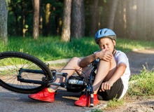 De jonge Kaukasische jongen in helm en witte t-shirt kreeg ongeval en zit op de grond na het vallen van de fiets en voelt pijn royalty-vrije stock afbeelding De jonge Kaukasische jongen in helm en witte t-shirt kreeg ongeval en zit op de grond na het vallen van de fiets en voelt pijn royalty-vrije stock afbeelding