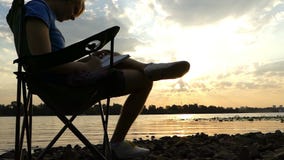 De jonge mens zit, denkt, schrijft iets in zijn blocnote, op een riverbank stock videobeelden De jonge mens zit, denkt, schrijft iets in zijn blocnote, op een riverbank stock videobeelden