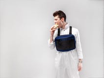 De jonge modieuze mannelijke arbeider met de zak van de borstinstallatie eet een smakelijke sappige hamburger op een witte achter stock afbeeldingen De jonge modieuze mannelijke arbeider met de zak van de borstinstallatie eet een smakelijke sappige hamburger op een witte achter stock afbeeldingen