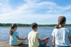 De jongen en twee meisjes zitten door het meer en doen yoga stock afbeelding De jongen en twee meisjes zitten door het meer en doen yoga stock afbeelding
