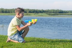 De jongen zit op het gras en schiet water van een pistool op het meer in de zomer stock foto's De jongen zit op het gras en schiet water van een pistool op het meer in de zomer stock foto's