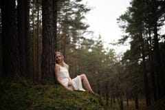 De mooie jonge zitting van de blondevrouw in bosnimf in witte kleding in altijdgroen hout stock foto's De mooie jonge zitting van de blondevrouw in bosnimf in witte kleding in altijdgroen hout stock foto's