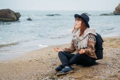 De mooie roodharige vrouw in een hoed en een sjaal met een rugzak zit in een meditatieve positie inzake de kust tegen stock afbeeldingen De mooie roodharige vrouw in een hoed en een sjaal met een rugzak zit in een meditatieve positie inzake de kust tegen stock afbeeldingen