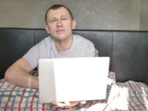 De slaperige jonge mens zit in bed, houdt laptop royalty-vrije stock fotografie De slaperige jonge mens zit in bed, houdt laptop royalty-vrije stock fotografie
