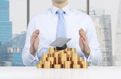De student plaatst zijn handen over de graduatiehoed en de muntstukkenpiramide Een concept een hoge prijs voor het universitaire royalty-vrije stock fotografie De student plaatst zijn handen over de graduatiehoed en de muntstukkenpiramide Een concept een hoge prijs voor het universitaire royalty-vrije stock fotografie
