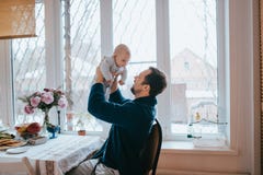 De vader houdt in zijn wapens zijn uiterst kleine zoonszitting op een stoel naast een groot venster in de keuken royalty-vrije stock afbeelding De vader houdt in zijn wapens zijn uiterst kleine zoonszitting op een stoel naast een groot venster in de keuken royalty-vrije stock afbeelding