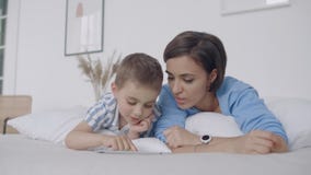 In de witte slaapkamer, bekijken het mamma en de zoon het de tabletscherm en lach Gelukkige familie in bed in de ochtend die een stock videobeelden In de witte slaapkamer, bekijken het mamma en de zoon het de tabletscherm en lach Gelukkige familie in bed in de ochtend die een stock videobeelden