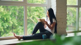 Den attraktiva asiatiska flickastudenten är läsebok- och lesammanträde på fönsteravsatsen i modern lägenhet Hobby ungdom lager videofilmer Den attraktiva asiatiska flickastudenten är läsebok- och lesammanträde på fönsteravsatsen i modern lägenhet Hobby ungdom lager videofilmer