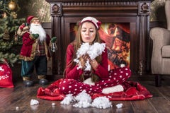 Den härliga le flickan i hem- kläderpyjamas för röd jul sitter under ett julträd nära en spis och blåser konstgjort s royaltyfria bilder Den härliga le flickan i hem- kläderpyjamas för röd jul sitter under ett julträd nära en spis och blåser konstgjort s royaltyfria bilder