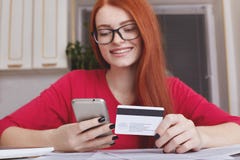 Den Redhaired nätta kvinnliga modellen i eyewearhåll ilar telefonen, och kreditkorten, gör online-köp eller shopping i internetla Arkivfoton Den Redhaired nätta kvinnliga modellen i eyewearhåll ilar telefonen, och kreditkorten, gör online-köp eller shopping i internetla Arkivfoton