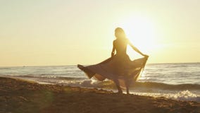 Den unga charmiga flickan i lång ljus klänning virvlar runt omkring på den sandiga stranden enlighted av guld- solnedgångstrålar arkivfilmer Den unga charmiga flickan i lång ljus klänning virvlar runt omkring på den sandiga stranden enlighted av guld- solnedgångstrålar arkivfilmer