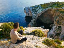 Den unga mannen sitter på kanten av en klippa och beundrar havet och stenarna royaltyfria foton Den unga mannen sitter på kanten av en klippa och beundrar havet och stenarna royaltyfria foton