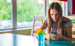 Den unga tonåringbrunettflickan med långt hår som inomhus sitter i stads- kafé, dricker en blå lemonadcoctail och använder hennes royaltyfria bilder Den unga tonåringbrunettflickan med långt hår som inomhus sitter i stads- kafé, dricker en blå lemonadcoctail och använder hennes royaltyfria bilder
