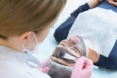 Der Cosmetologist in den rosa Handschuhen mit einer Bürste wendet eine Kohlenstoffmaske für die Schale auf das Gesicht eines jung lizenzfreies stockbild Der Cosmetologist in den rosa Handschuhen mit einer Bürste wendet eine Kohlenstoffmaske für die Schale auf das Gesicht eines jung lizenzfreies stockbild