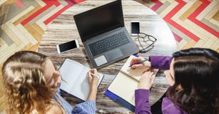 Deux jeunes femmes faisant des disques dans un carnet dans une vue supérieure de bureau Ordinateur portable, verres, téléphone in images stock Deux jeunes femmes faisant des disques dans un carnet dans une vue supérieure de bureau Ordinateur portable, verres, téléphone in images stock