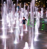 Die glücklichen Kinder, die in einem Wasserbrunnen am Abend spielen, beleuchtet lizenzfreies stockfoto Die glücklichen Kinder, die in einem Wasserbrunnen am Abend spielen, beleuchtet lizenzfreies stockfoto