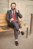 Directeur de sourire s'asseyant au banc et à téléphoner Jeune emplacement souri bel d'homme d'affaires sur le banc avec son ordin image stock Directeur de sourire s'asseyant au banc et à téléphoner Jeune emplacement souri bel d'homme d'affaires sur le banc avec son ordin image stock