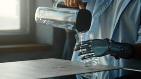 Disabled Guy With Bio Hand Prothesis Pours Water Into Glass From Bowl And Drinks Standing In Kitchen Stock Photography