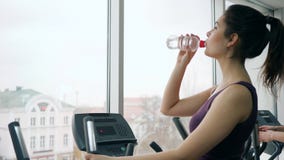 Dissetante nella palestra, ragazza funziona sul simulatore di sport e beve l'acqua dalla bottiglia stock footage Dissetante nella palestra, ragazza funziona sul simulatore di sport e beve l'acqua dalla bottiglia stock footage