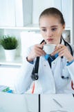 Doctor caucásico de sexo femenino joven que tiene una rotura y que bebe el café que se sienta en su lugar de trabajo imágenes de archivo libres de regalías Doctor caucásico de sexo femenino joven que tiene una rotura y que bebe el café que se sienta en su lugar de trabajo imágenes de archivo libres de regalías