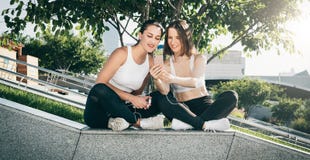 Dois atletas das jovens mulheres no sportswear estão sentando-se no parque, relaxam após esportes treinando, usam o smartphone imagem de stock Dois atletas das jovens mulheres no sportswear estão sentando-se no parque, relaxam após esportes treinando, usam o smartphone imagem de stock