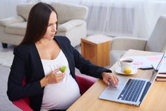 Donna di affari incinta seria che lavora al computer portatile e che mangia maca immagini stock libere da diritti Donna di affari incinta seria che lavora al computer portatile e che mangia maca immagini stock libere da diritti