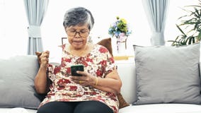 Donna senior asiatica che per mezzo dello Smart Phone e bevendo una tazza di caffè a casa archivi video Donna senior asiatica che per mezzo dello Smart Phone e bevendo una tazza di caffè a casa archivi video