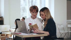 Dos encargados de sexo femenino jovenes hermosos que trabajan así como el ordenador portátil y la libreta, discuten el trabajo en metrajes Dos encargados de sexo femenino jovenes hermosos que trabajan así como el ordenador portátil y la libreta, discuten el trabajo en metrajes