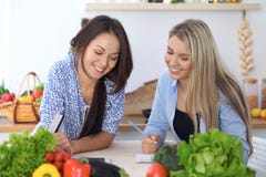Dos mujeres felices jovenes que hacen las compras en línea para hacer el menú por la tableta Amigos que cocinan en la cocina imagen de archivo Dos mujeres felices jovenes que hacen las compras en línea para hacer el menú por la tableta Amigos que cocinan en la cocina imagen de archivo