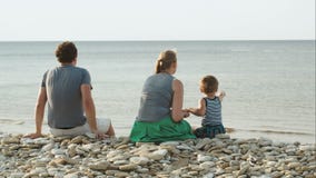 Dreiköpfige Familie, die auf Pebble Beach durch sitzt stock video footage Dreiköpfige Familie, die auf Pebble Beach durch sitzt stock video footage