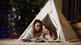 Dressed in white sweaters, my mother and daughter read a fairy tale for the night lying with her in the tent of an stock video footage Dressed in white sweaters, my mother and daughter read a fairy tale for the night lying with her in the tent of an stock video footage