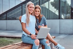 Dwa pięknej dziewczyny siedzi chwyty i patrzeje kamerę laptop podczas gdy siedzący na ławce na tle obrazy stock Dwa pięknej dziewczyny siedzi chwyty i patrzeje kamerę laptop podczas gdy siedzący na ławce na tle obrazy stock