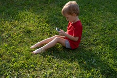Dziecko chłopiec spojrzenia w telefon komórkowego podczas gdy siedzący na trawie Pojęcie edukacja i zależność na gadżetach w dzie obraz stock Dziecko chłopiec spojrzenia w telefon komórkowego podczas gdy siedzący na trawie Pojęcie edukacja i zależność na gadżetach w dzie obraz stock