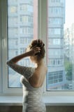 Dziewczyna z długim brązu włosy w białej smokingowej pozycji przy okno od plecy i koryguje fryzurę fotografia royalty free Dziewczyna z długim brązu włosy w białej smokingowej pozycji przy okno od plecy i koryguje fryzurę fotografia royalty free