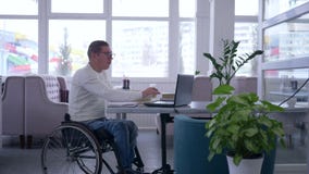 E-lerend, lezen de hogere mannelijke gehandicapten in rolstoel die glazen dragen boek en gebruiken laptop zitting bij een lijst i stock footage E-lerend, lezen de hogere mannelijke gehandicapten in rolstoel die glazen dragen boek en gebruiken laptop zitting bij een lijst i stock footage