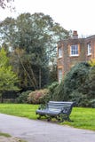 Een bank in het park langs de weg tegen de achtergrond van een klassiek Engels rood baksteengebouw royalty-vrije stock fotografie Een bank in het park langs de weg tegen de achtergrond van een klassiek Engels rood baksteengebouw royalty-vrije stock fotografie