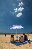 Een groep die mensen onder een blauwe en witte strandparaplu tegen een heldere blauwe hemel zitten Stock Fotografie Een groep die mensen onder een blauwe en witte strandparaplu tegen een heldere blauwe hemel zitten Stock Fotografie