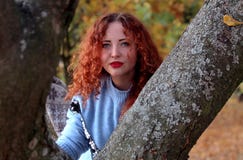 Een jonge vrouw met rood haar onderzoekt aandachtig de camera en klampt zich aan een boom in het park vast op de achtergrond kunt stock foto Een jonge vrouw met rood haar onderzoekt aandachtig de camera en klampt zich aan een boom in het park vast op de achtergrond kunt stock foto