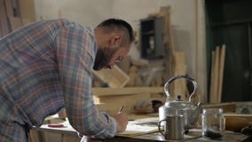 Een mens in een timmerwerkworkshop maakt berekeningen bij de lijst stock footage Een mens in een timmerwerkworkshop maakt berekeningen bij de lijst stock footage