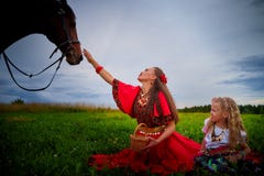 image photo : A woman and a girl in bright Gypsy dresses with a horse in a field with green grass. Mother and daughter pose in nature with an