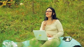 Een vrouw met glazen op de rand van een bos met een potlood in haar hand en laptop denkt over het project stock video Een vrouw met glazen op de rand van een bos met een potlood in haar hand en laptop denkt over het project stock video