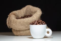 Een witte koffiemok met koffiebonen binnen en een koffiezak op witte lijst en zwarte achtergrond royalty-vrije stock afbeeldingen Een witte koffiemok met koffiebonen binnen en een koffiezak op witte lijst en zwarte achtergrond royalty-vrije stock afbeeldingen