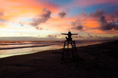 Een zitting van de silhouetvrouw op een badmeestertoren met open wapens die van de zonsopgang op een strand en de zon` s stralen stock afbeelding Een zitting van de silhouetvrouw op een badmeestertoren met open wapens die van de zonsopgang op een strand en de zon` s stralen stock afbeelding