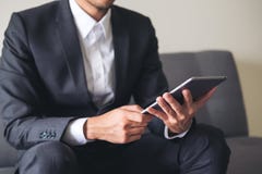 Ein Geschäftsmann in einer grauen Klage, die auf Sofaholding sitzt, Tabletten-PC nach Arbeit verwendet und betrachtet lizenzfreie stockbilder Ein Geschäftsmann in einer grauen Klage, die auf Sofaholding sitzt, Tabletten-PC nach Arbeit verwendet und betrachtet lizenzfreie stockbilder