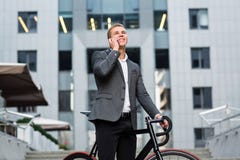 Ein junger Geschäftsmann, der auf den Schritten eines Bürogebäudes, mit einem Ordner von Papieren und von Fahrrad steht Junges hü stockbild Ein junger Geschäftsmann, der auf den Schritten eines Bürogebäudes, mit einem Ordner von Papieren und von Fahrrad steht Junges hü stockbild